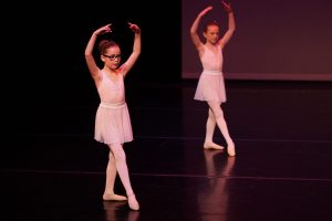 Two elementary aged dance students performing in dance recital in pose with arms curved overhead and one foot pointed to the front.