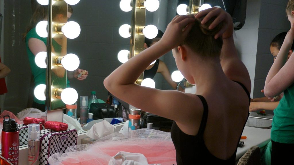 Young dance students backstage putting hair in bun for showcase performance.