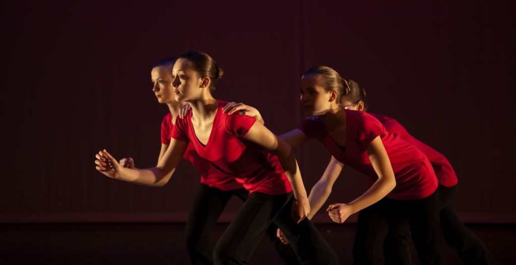 Group of four teen dancers at Leica Hardy School of Dance bent forward as if running, holding shoulders, looking into stage light.