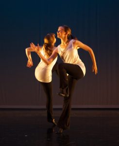 Two teenaged girls dancing energetically facing opposite directions on the diagonal. They are standing on one leg with the other leg folded up in front. Their arms are posed as if they are running.