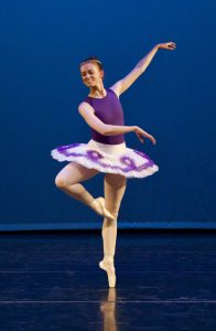 Photo of ballet dancer dressed in purple tutu on pointe. One leg is bent with the toe touching the knee of the standing leg.