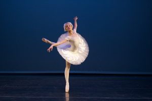 Solo dancer on pointe performing Swan Lake variation in recital. She is facing the camera with arms outstretched in wide diagonal with one leg reaching up to the back.