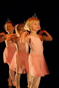 Three Grade Primary ballet students walking on toes with hands lightly placed on shoulders like wings. They are dressed in pink chiffon skirts and sliver crowns.