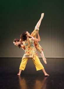 Two contemporary dancers partnering each other. One dancer in a broad stance with her back to the audience lifts the other dancer who is tipping forward holding her legs in the splits.