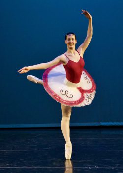 Solo dancer on pointe on her right leg with the left leg curved backwards at waist height bent at the knee. Her left arm is overhead in a long arc and her right arm is outstretched softly to the side at shoulder height.