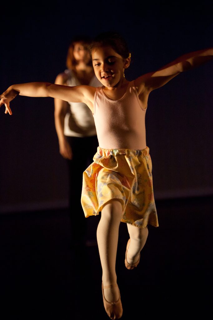 Excited smiling child in a dance recital. She is leaping with both legs bent. Her arms are thown wide open.