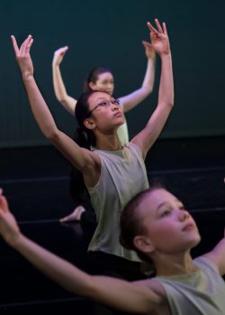 Three young teenaged dancers in a row. Their arms are overhead and U-shaped. They are lifting their chests in a slight back arch as they gaze diagonally upward.