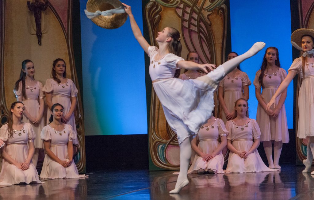 Photo from the Nutcracker. A solo dancer is joyously holding a hat in front of her. She is standing on the balls of one foot with the other leg tossed up behind her at shoulder height. 