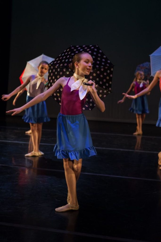 Group of teenaged dancers in recital. They are posed holding umbrellas over one shoulder. Their heads are turned to one side with the opposite softly held outward.