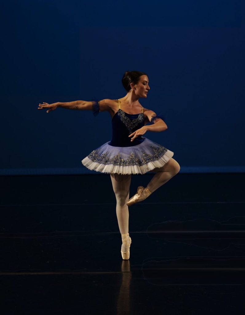Dancer in pointe shoes standing on one leg with other leg bent at the knee. The toe of the bent leg is touching the knee of the standing leg. Her arms are swept across her body in the opposite direction of the knee. She is regally gazing downward towards the bent knee.