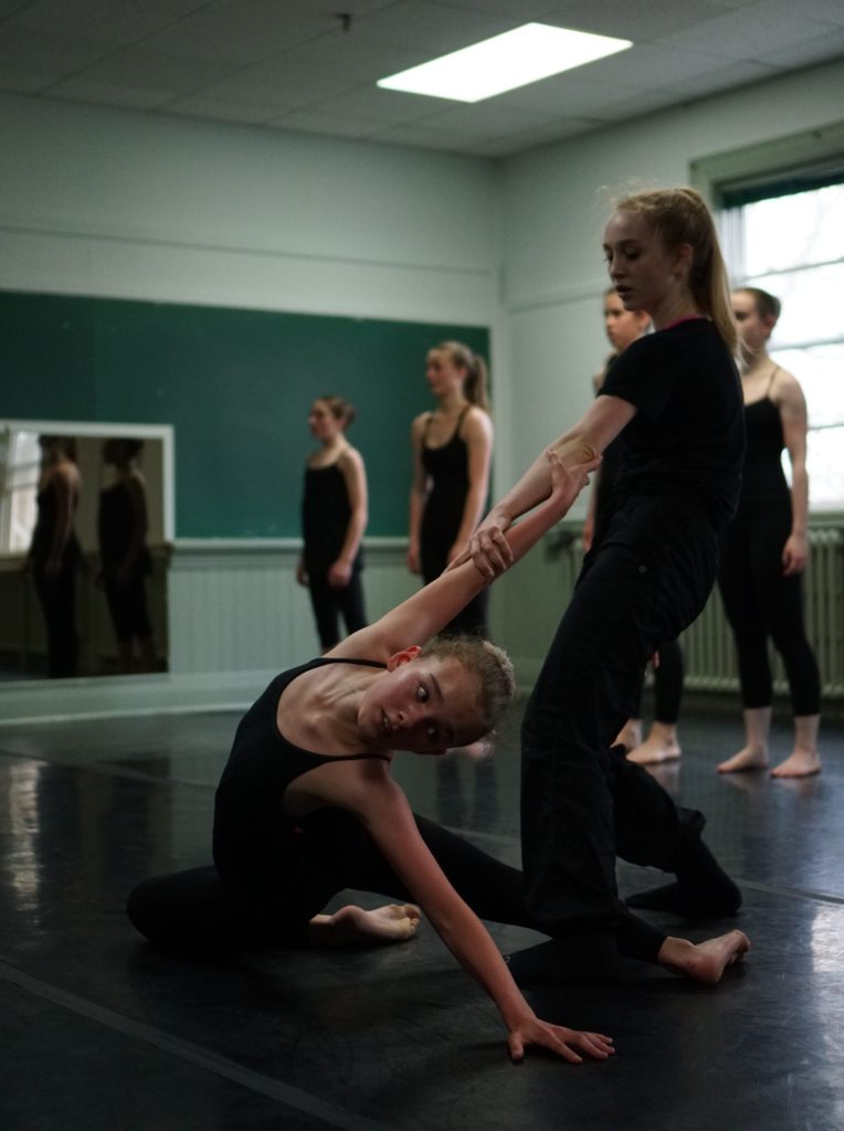 Photo of a group of dancers in rehearsal. The two dancers in the foreground are partnering each other by clasping wrists. The standing dancer is holding the arm of the other as she leans slightly backward to counterbalance the weight of the other. The other dancer is paritially seated on the floor with both legs bent, one in front and the other behind. She is arching deeply backwards while reaching backwards with her free arm. 