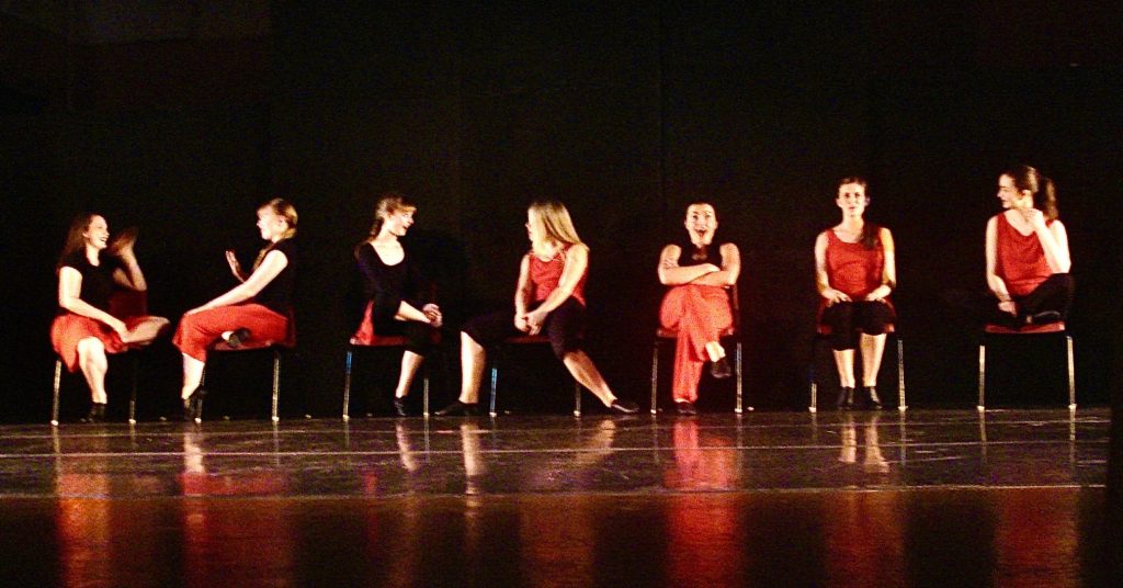 Group of seven dancers sitting in a row of chairs stretched across the stage width-wise. They are in animated conversational poses. 