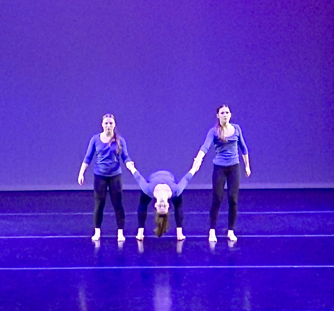 A trio of dancers are holding hands stretched across the stage. The centre dancer's body is facing backwards to the audience. She is in a very deep backbend looking a the audience with her head almost touching the floor. 
