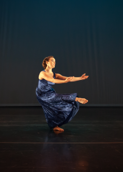 Photo of a solo modern dancer. She is dressed in a royal blue long velvet dress. She is standing on a single leg, bent at the knee and leaning backwards off-centre. Both of her arms are reaching forward as if offering something to the viewer. Her other leg is also reaching forward horizonally to the floor.