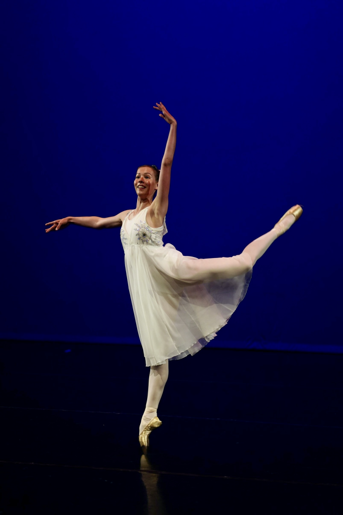 Photo of solo dancer in white chiffon dress. She is standing on one leg on pointe with her other leg extended backwards bent arching upwards at the knee. One arm is curved overhead and the other arm is stretched sideways parallel to the floor.