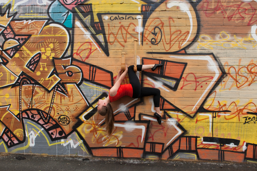 Photo of dancer posing in front a a wall of grafitti that can be found on the Dartmouth Harbourwalk Trail. She is hanging upside down from a pipe protruding from the wall. She is arching backwards with her legs bent at the knee. She is wearing black leggings and an orange T-shirt, which makes her appear to be part of the graffiti. 