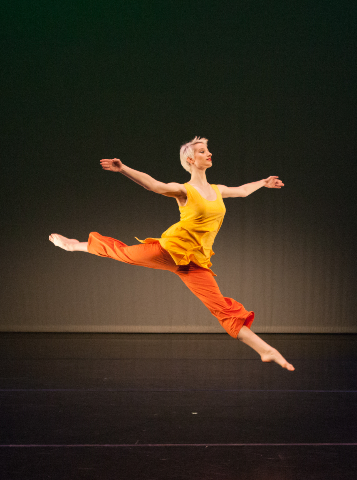 Photo of a solo modern dancer. She is leapingin the air with her legs in a split. Her arms are stetched sideways with palms facing forwards and chest arching upwards.