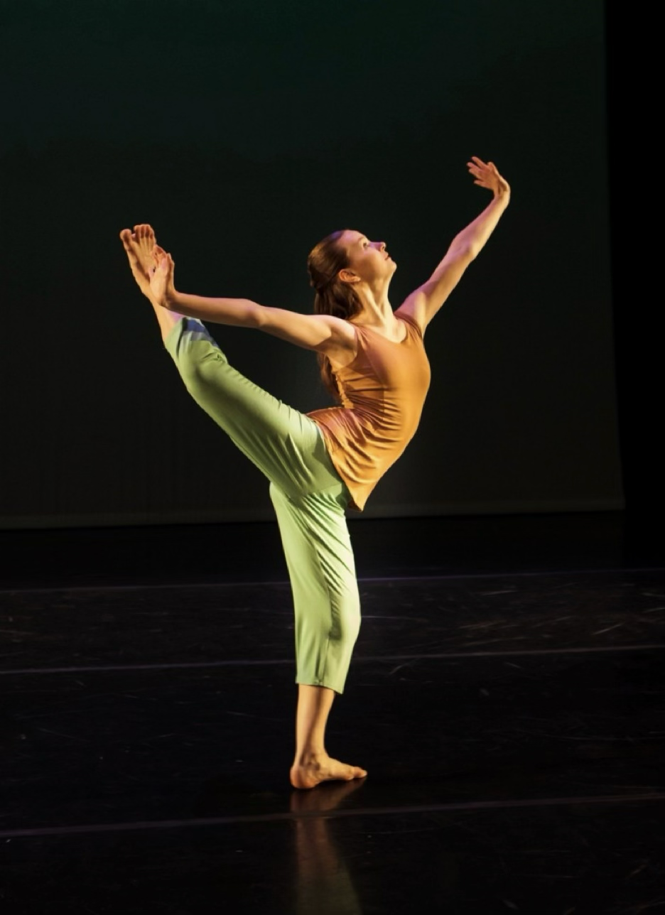 Photo of solo modern dancer. She is standing on a bent leg. The other leg is extended backwards bent at the knee at head height. She is arching her back looking upwards. Her arms are outstretched with flexed wrists.