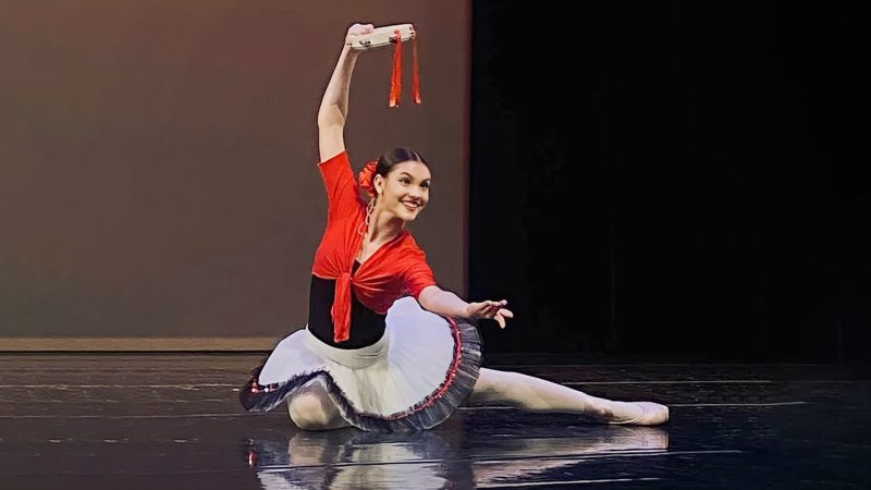 Smiling dancer dressed in Spanish-styled tutu posed kneeling on floor holding tambourine.