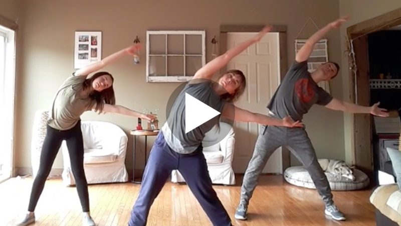 Photo of a mother, father and young girl exercising together. They arranged in a triangle with the mother leading the workout class. They are leaning to their left with their legs apart in a wide stance and their arms reaching in a "V" sideways.