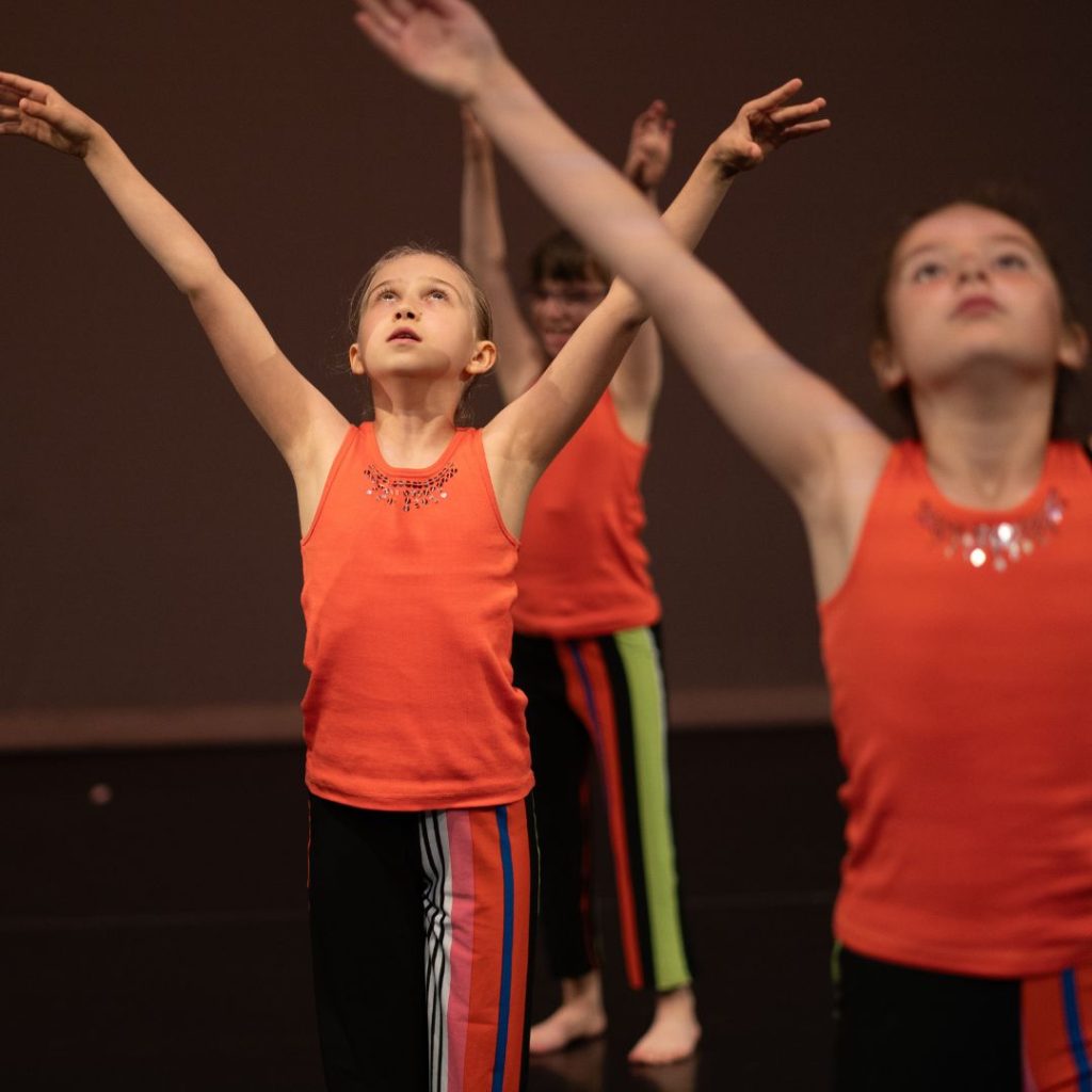 Young dancers performing on stage in orange tops and striped pants with arms raised during a contemporary dance performance.