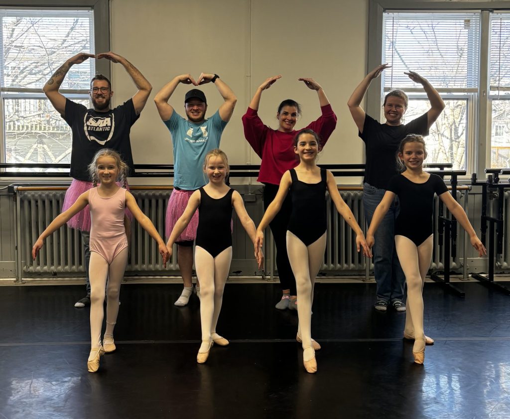 A joyful ballet class featuring young dancers in leotards and tights posing gracefully in front of smiling adults wearing casual clothes and tutus. The group stands in a bright dance studio with large windows, showcasing teamwork, fun, and the joy of dance education.